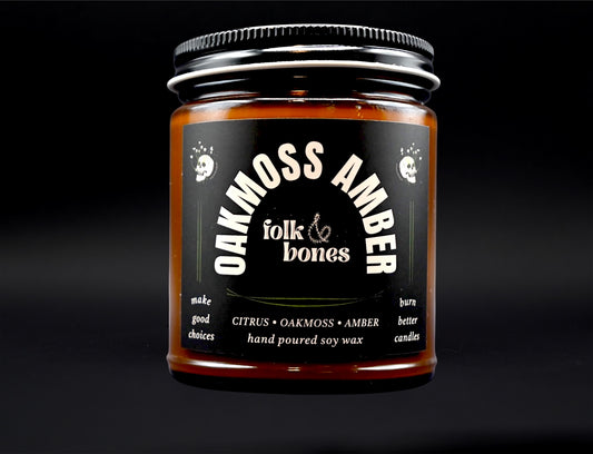 Folk and Bones Oakmoss Amber soy candle in an 8 oz glass jar with a lit wick, showcasing a cozy, earthy ambiance.