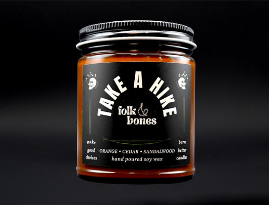 Folk and Bones Take a Hike 8 oz soy candle with a rustic label and natural wood wick, perfect for cozy home decor.