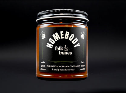 Folk and Bones Homebody 8 oz soy candle in a glass jar with a wooden lid, warm amber glow and clean-burning wick.