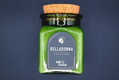 Folk and Bones Belladonna 8 oz soy candle in a glass jar with a lit wick and decorative label, ideal for home ambiance.