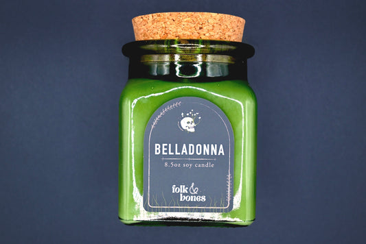 Folk and Bones Belladonna 8 oz soy candle in a glass jar with a lit wick and decorative label, ideal for home ambiance.