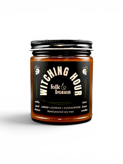 Folk and Bones Witching Hour 8oz soy candle in a dark jar with a black lid, featuring a mystical label and a lit wick.